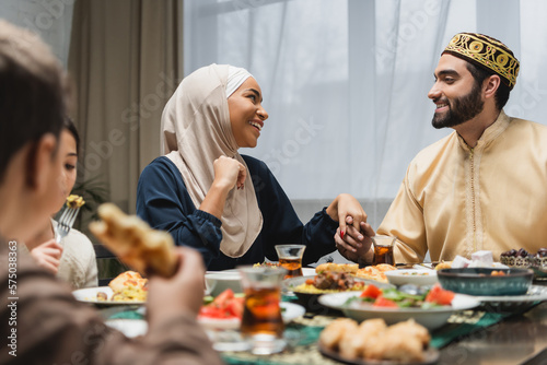 Smiling muslim family holding hands near kids and ramadan dinner.