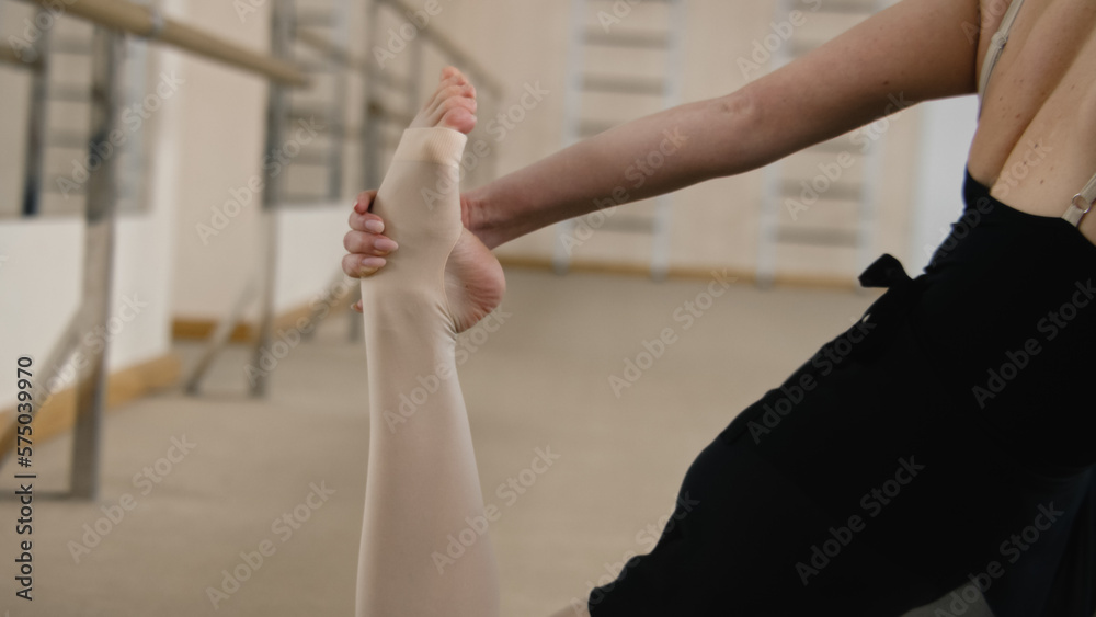 Ballerina in training bodysuit stands near ballet barre in dance studio, stretches her leg and prepares for performance. Female ballet dancer doing gymnastic exercises. Classical ballet dance school.