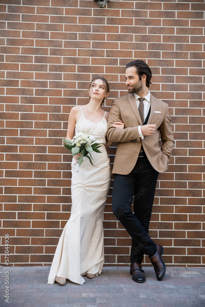 full length of dreamy bride in wedding dress holding flowers near groom in suit while standing against brick wall. full length of dreamy bride in wedding dress holding flowers near groom in suit while standing against brick wall.