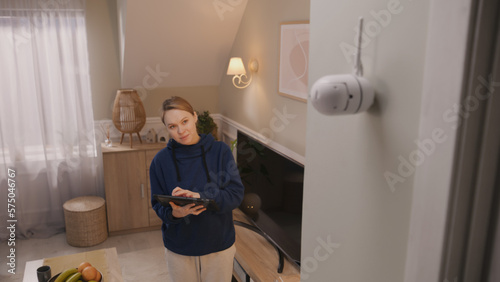 Caucasian woman installs security camera. Woman sets up angle of CCTV camera at home and rotates it with digital tablet computer and application. Concept of monitoring system, safety and privacy.