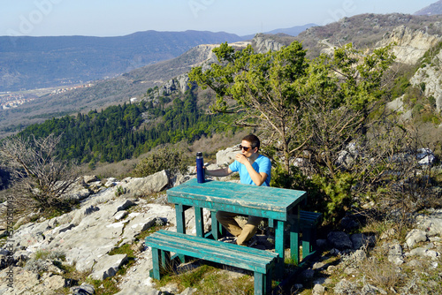 A tourist drinks a drink from a thermos, sitting on a bench in a picturesque mountainous area - a portrait of a man taken during outdoor recreation.