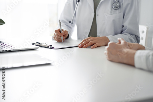 Doctor and patient sitting at the desk in clinic office. The focus is on female physician's hands filling up the medication history record form, close up. Perfect medical service and medicine concept.