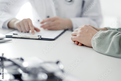 Doctor and patient sitting at the desk in clinic office. The focus is on female physician's hands filling up the medication history record form, close up. Medicine concept.