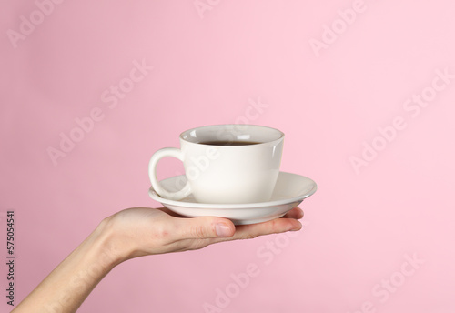 Female hand hold a white ceramic cup with a saucer on a pink background