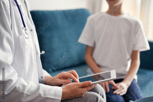 Doctor woman and kid boy patient at home. The pediatrician using tablet computer while filling up medical records, close up. Medicine, healthcare concepts.