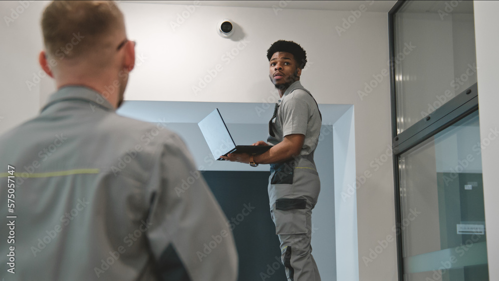 Two multi ethnic installers in uniform sets up security camera in office hallway using laptop. They talk, check CCTV cameras in computer program. Concept of surveillance system and privacy. Dolly shot Two multi ethnic installers in uniform sets up security camera in office hallway using laptop. They talk, check CCTV cameras in computer program. Concept of surveillance system and privacy. Dolly shot