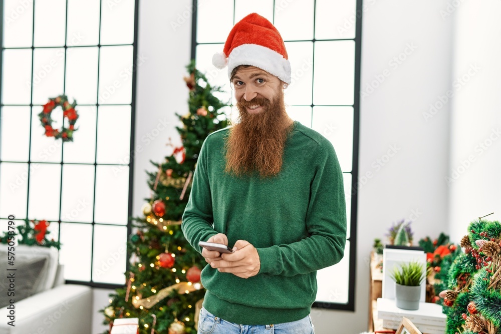 Young redhead man using smartphone standing by christmas tree at home Young redhead man using smartphone standing by christmas tree at home