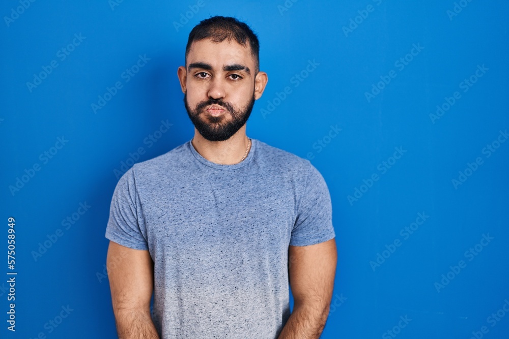 Middle east man with beard standing over blue background puffing cheeks with funny face. mouth inflated with air, crazy expression. Middle east man with beard standing over blue background puffing cheeks with funny face. mouth inflated with air, crazy expression.