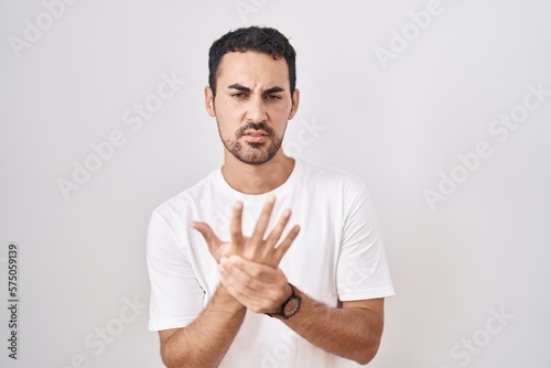 Handsome hispanic man standing over white background suffering pain on hands and fingers, arthritis inflammation