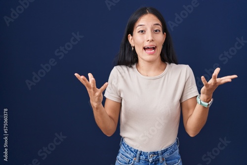 Young hispanic woman standing over blue background celebrating crazy and amazed for success with arms raised and open eyes screaming excited. winner concept