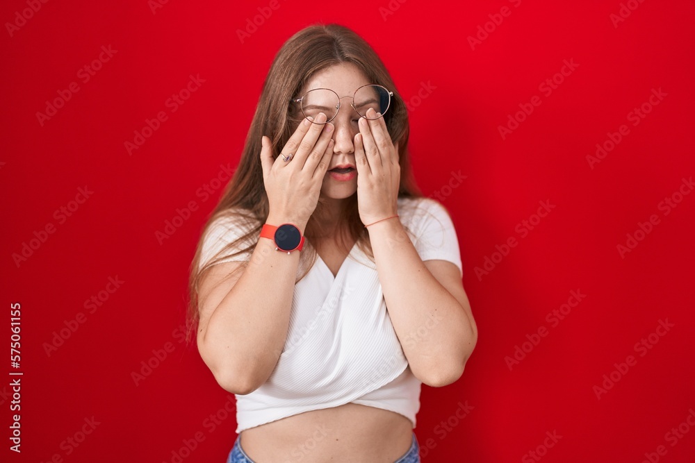 Young caucasian woman standing over red background rubbing eyes for fatigue and headache, sleepy and tired expression. vision problem Young caucasian woman standing over red background rubbing eyes for fatigue and headache, sleepy and tired expression. vision problem
