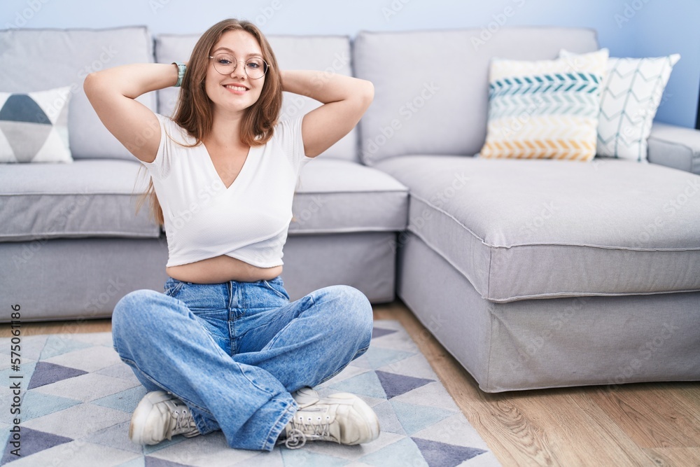 Young caucasian woman sitting on the floor at the living room relaxing and stretching, arms and hands behind head and neck smiling happy