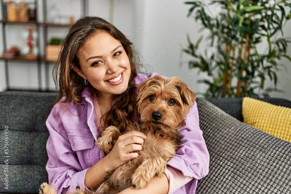 Young beautiful hispanic woman hugging dog sitting on sofa at home Young beautiful hispanic woman hugging dog sitting on sofa at home