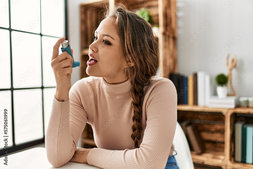 Young beautiful hispanic woman using inhaler sitting on table at home - obrazy, fototapety, plakaty 