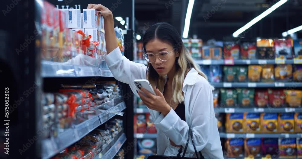 Portrait of asian female buyer looks into smartphone and then chooses a brand of food products for purchase on a shelf with food. Asian woman buyer with smartphone buying food in grocery supermarket