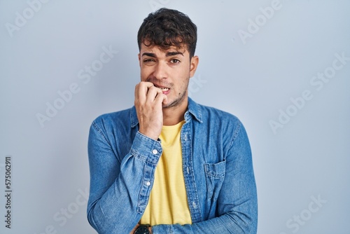 Young hispanic man standing over blue background looking stressed and nervous with hands on mouth biting nails. anxiety problem.