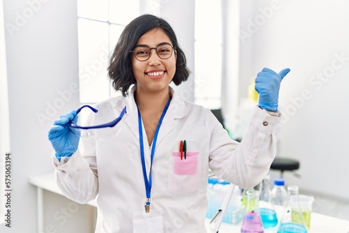 Young hispanic woman working at scientist laboratory holding safety glasses pointing thumb up to the side smiling happy with open mouth