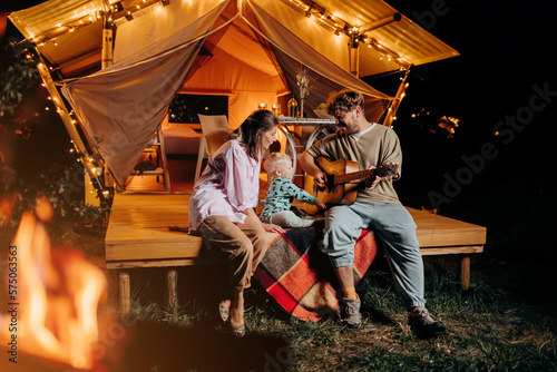 Happy family relaxing and spend time together in glamping on summer evening and playing guitar near cozy bonfire. Luxury camping tent for outdoor recreation and recreation. Lifestyle concept © bondvit