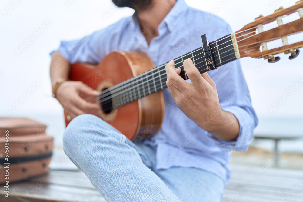 Young hispanic man musician playing classical guitar sitting on bench at seaside