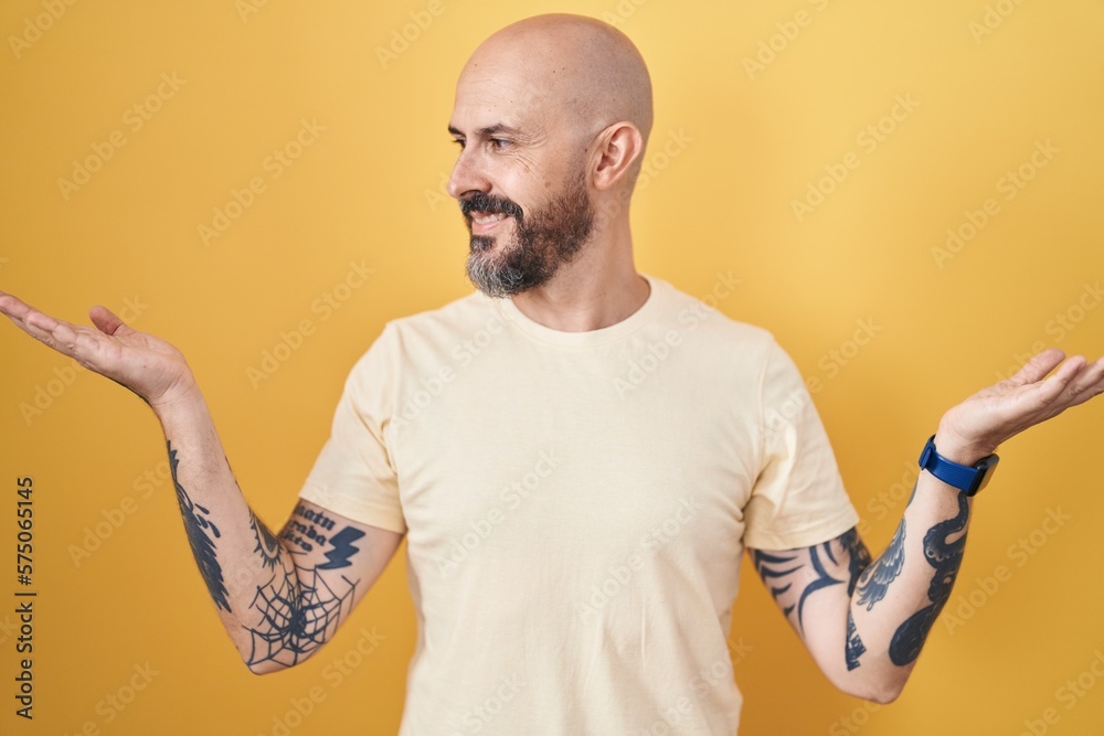 Hispanic man with tattoos standing over yellow background smiling showing both hands open palms, presenting and advertising comparison and balance