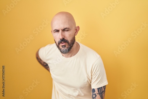 Hispanic man with tattoos standing over yellow background suffering of backache, touching back with hand, muscular pain