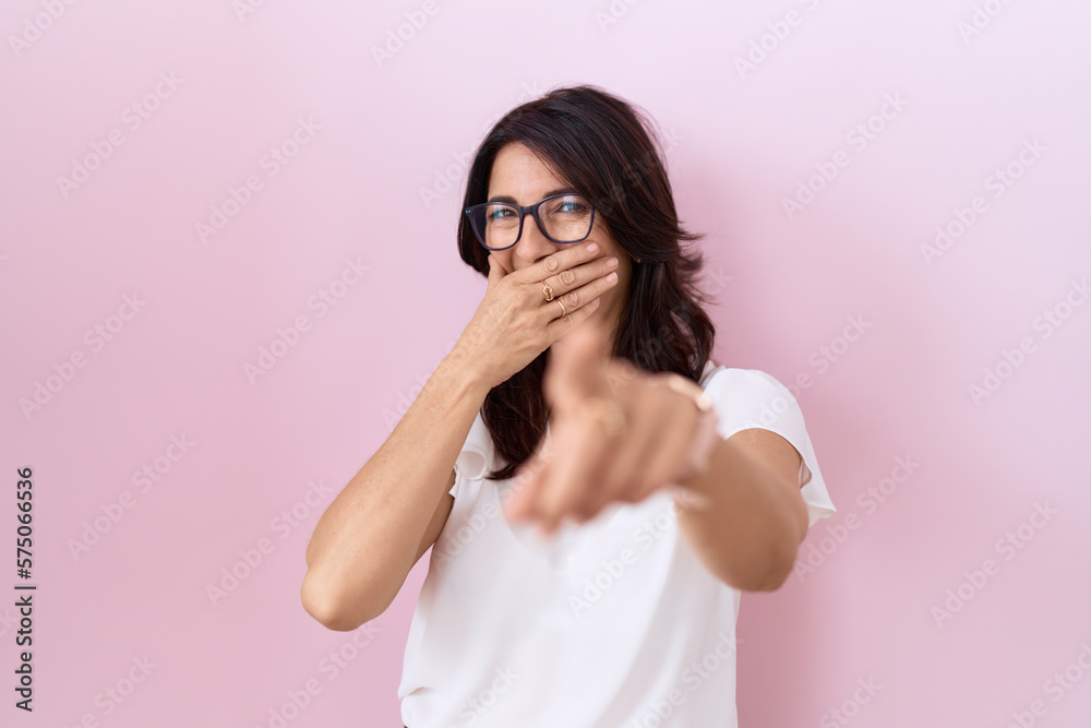 Middle age hispanic woman wearing casual white t shirt and glasses laughing at you, pointing finger to the camera with hand over mouth, shame expression Middle age hispanic woman wearing casual white t shirt and glasses laughing at you, pointing finger to the camera with hand over mouth, shame expression