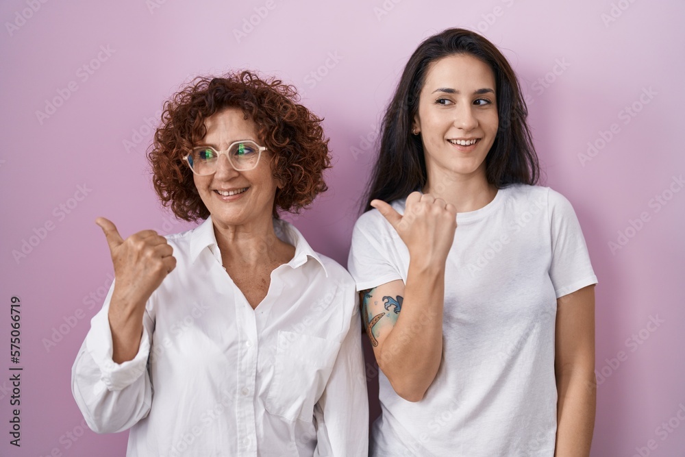 Hispanic mother and daughter wearing casual white t shirt over pink background smiling with happy face looking and pointing to the side with thumb up.