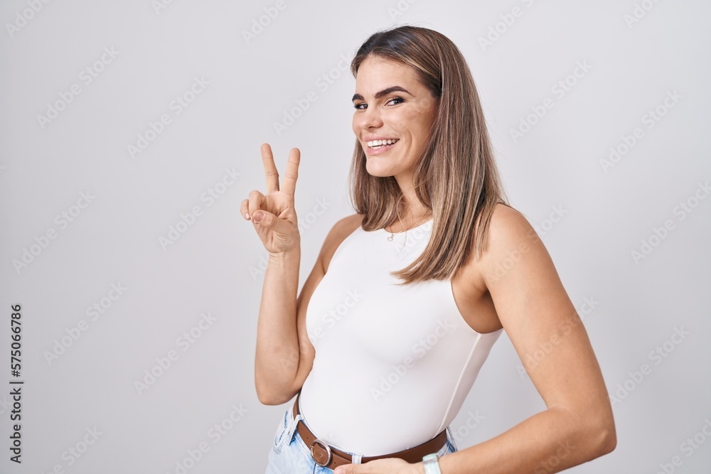 Hispanic young woman standing over white background smiling looking to the camera showing fingers doing victory sign. number two. Hispanic young woman standing over white background smiling looking to the camera showing fingers doing victory sign. number two.