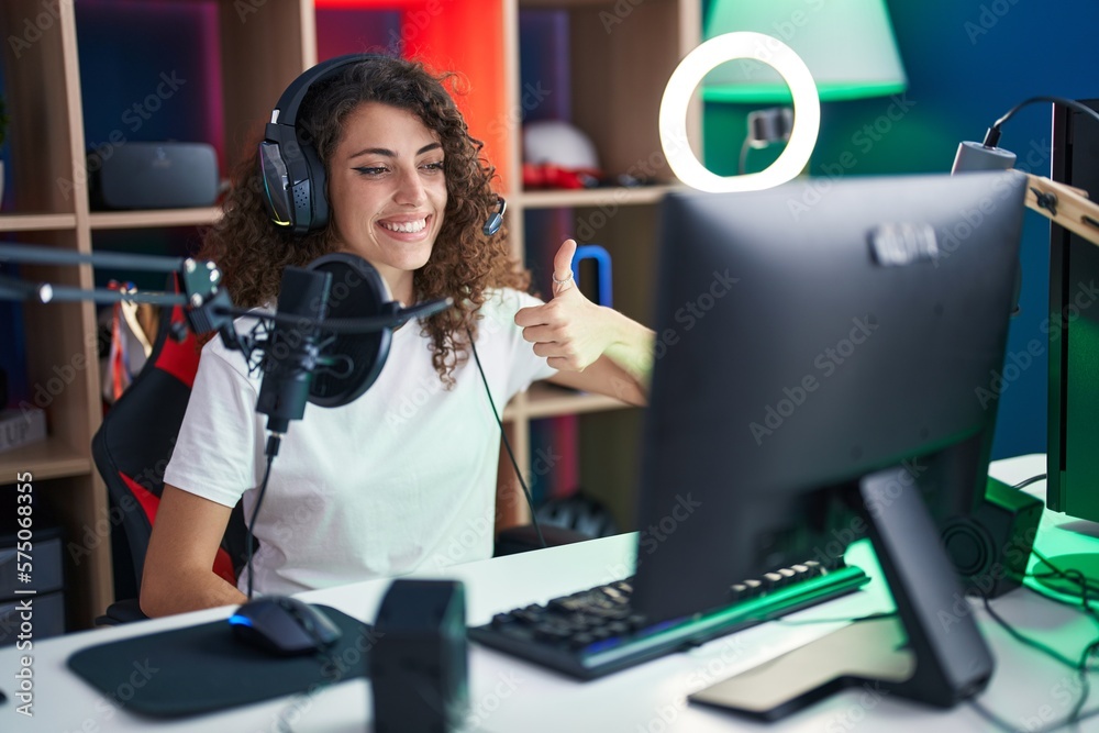 Hispanic woman with curly hair playing video games smiling happy and positive, thumb up doing excellent and approval sign