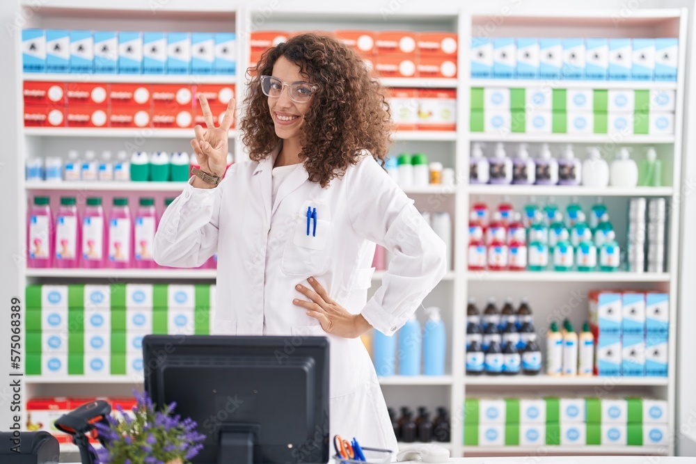 Hispanic woman with curly hair working at pharmacy drugstore smiling looking to the camera showing fingers doing victory sign. number two.