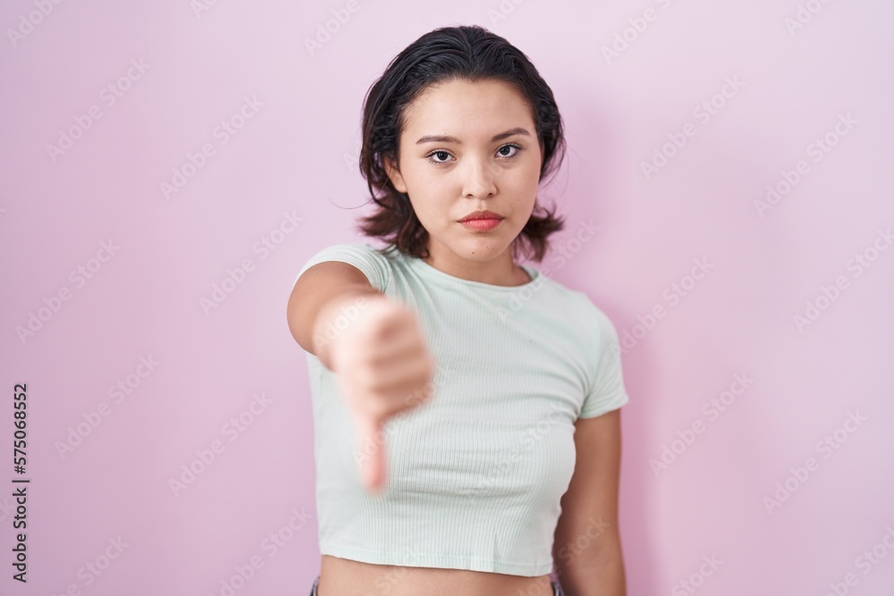 Hispanic young woman standing over pink background looking unhappy and angry showing rejection and negative with thumbs down gesture. bad expression.