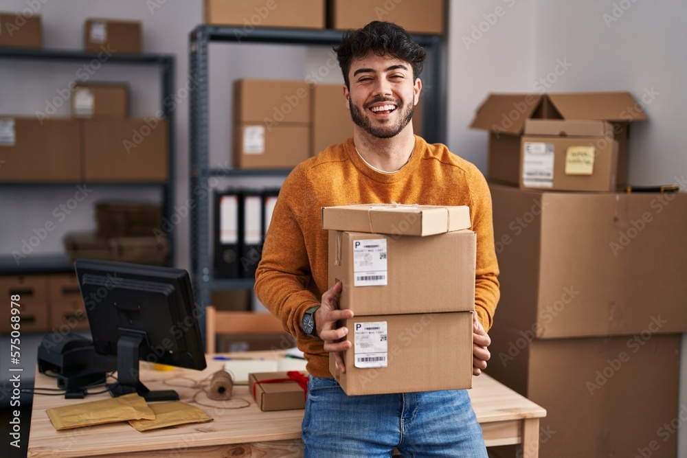 Hispanic man with beard working at small business ecommerce holding packages celebrating crazy and amazed for success with open eyes screaming excited. - obrazy, fototapety, plakaty 
