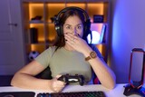 Beautiful brunette woman playing video games wearing headphones shocked covering mouth with hands for mistake. secret concept.