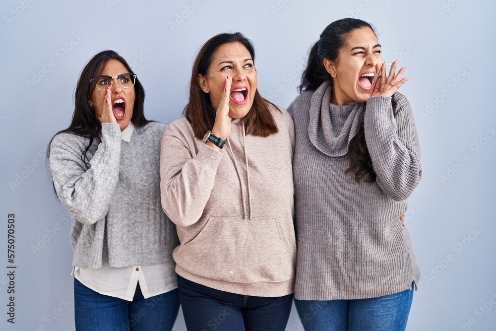 Mother and two daughters standing over blue background shouting and screaming loud to side with hand on mouth. communication concept. Mother and two daughters standing over blue background shouting and screaming loud to side with hand on mouth. communication concept.