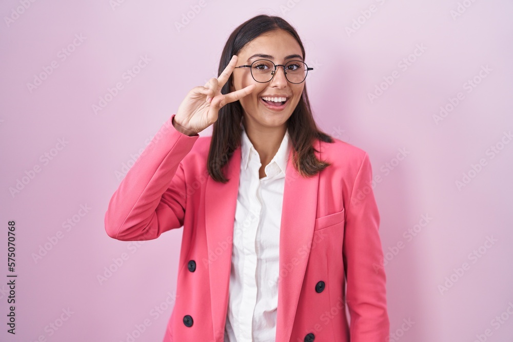 Young hispanic woman wearing business clothes and glasses doing peace symbol with fingers over face, smiling cheerful showing victory