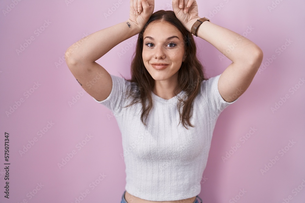 Young hispanic girl standing over pink background doing funny gesture with finger over head as bull horns Young hispanic girl standing over pink background doing funny gesture with finger over head as bull horns