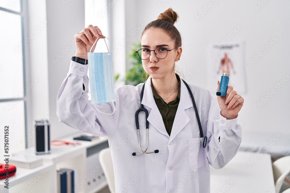Young hispanic girl holding medical asthma inhaler at the clinic puffing cheeks with funny face. mouth inflated with air, catching air. Young hispanic girl holding medical asthma inhaler at the clinic puffing cheeks with funny face. mouth inflated with air, catching air.