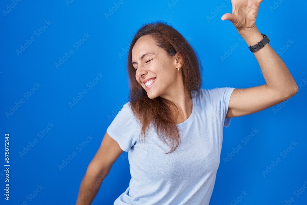 Fototapeta premium Brunette woman standing over blue background dancing happy and cheerful, smiling moving casual and confident listening to music