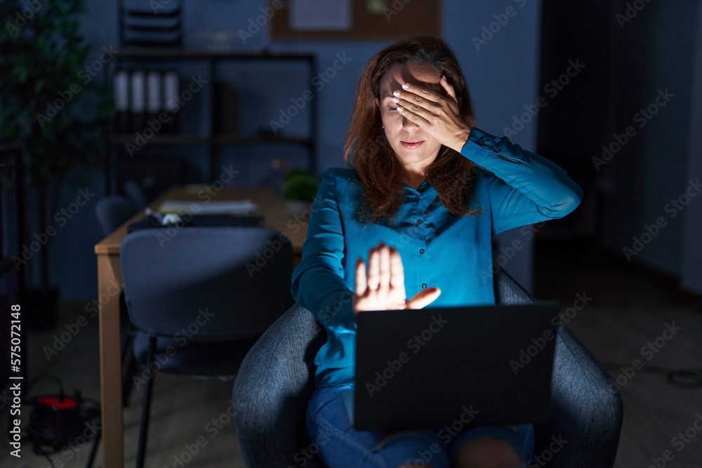 Brunette woman working at the office at night covering eyes with hands and doing stop gesture with sad and fear expression. embarrassed and negative concept. Brunette woman working at the office at night covering eyes with hands and doing stop gesture with sad and fear expression. embarrassed and negative concept.