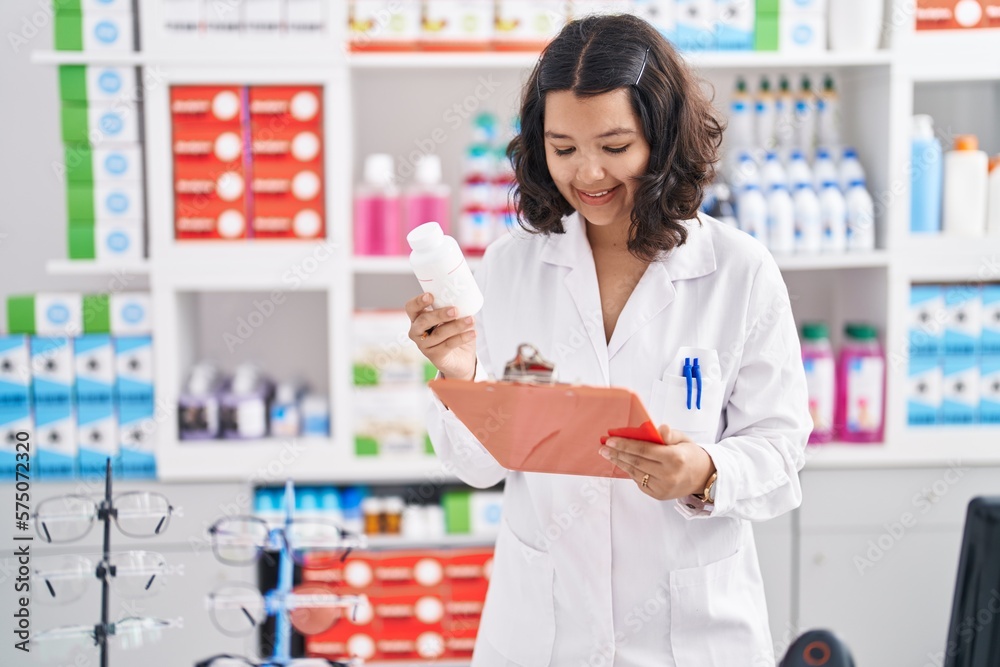 Young woman pharmacist holding pills bottle reading document at pharmacy
