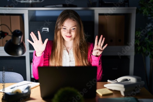Young caucasian woman working at the office at night showing and pointing up with fingers number nine while smiling confident and happy.
