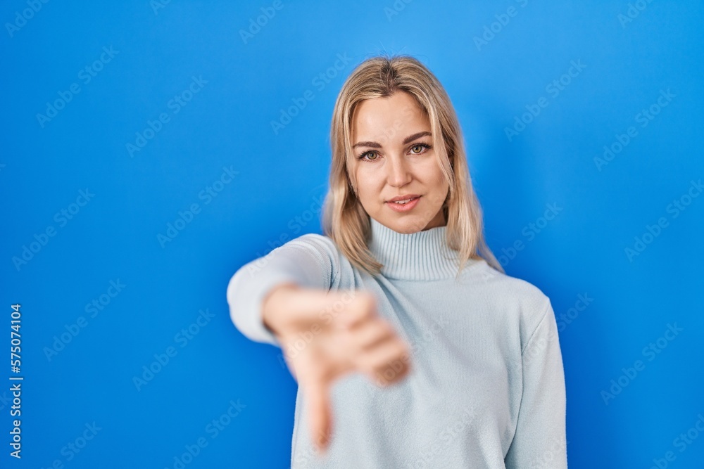 Fototapeta premium Young caucasian woman standing over blue background looking unhappy and angry showing rejection and negative with thumbs down gesture. bad expression.