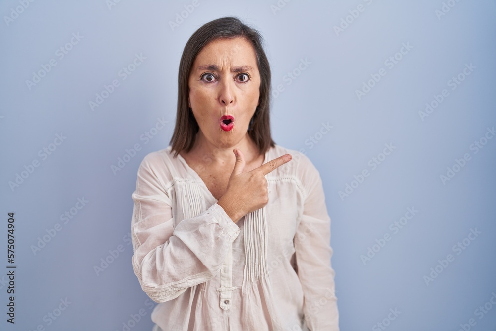 Middle age hispanic woman standing over blue background surprised pointing with finger to the side, open mouth amazed expression. Middle age hispanic woman standing over blue background surprised pointing with finger to the side, open mouth amazed expression.