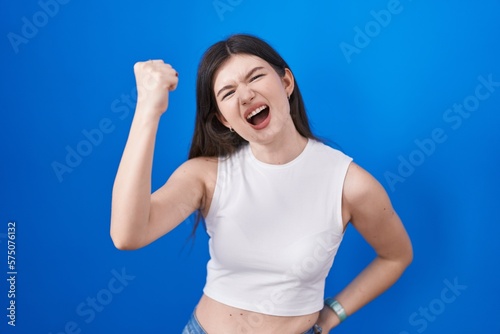 Young caucasian woman standing over blue background angry and mad raising fist frustrated and furious while shouting with anger. rage and aggressive concept.