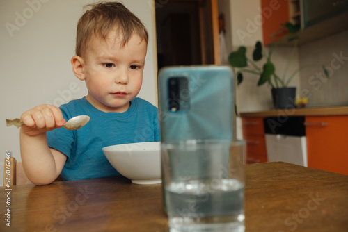 Child, a boy of three years old, is having breakfast at the kitchen table and watching cartoons on his smartphone