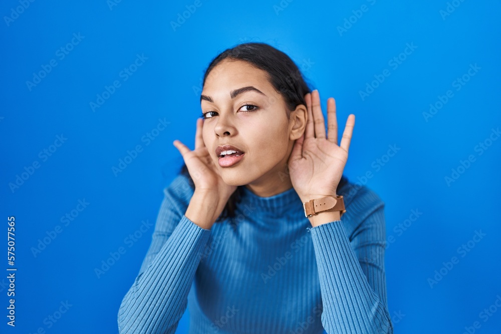 Young brazilian woman standing over blue isolated background trying to hear both hands on ear gesture, curious for gossip. hearing problem, deaf Young brazilian woman standing over blue isolated background trying to hear both hands on ear gesture, curious for gossip. hearing problem, deaf