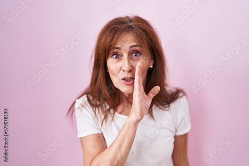 Middle age woman standing over pink background hand on mouth telling secret rumor, whispering malicious talk conversation