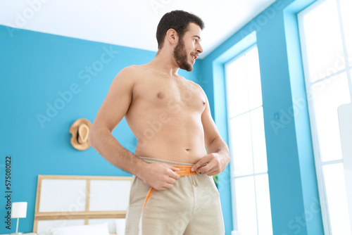 Young hispanic man measuring waist with tape measure at home