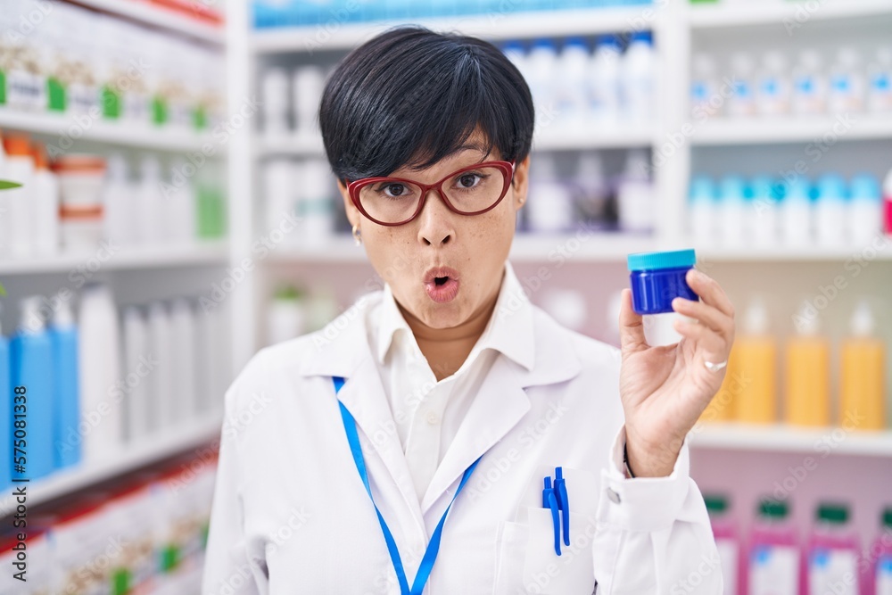 Young asian woman with short hair working at pharmacy drugstore holding cream jar scared and amazed with open mouth for surprise, disbelief face