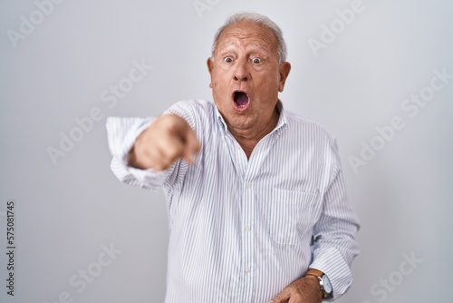 Senior man with grey hair standing over isolated background pointing with finger surprised ahead, open mouth amazed expression, something on the front